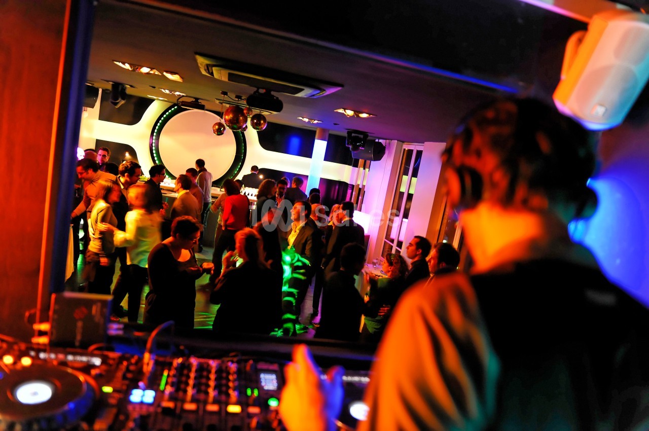 Un DJ mixe de la musique dans une salle animée où des personnes dansent et discutent sous des lumières colorées.