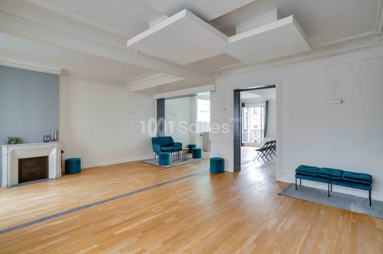 Salon spacieux avec parquet clair, cheminée, murs blancs, fauteuils bleus et ouverture sur une autre pièce lumineuse.