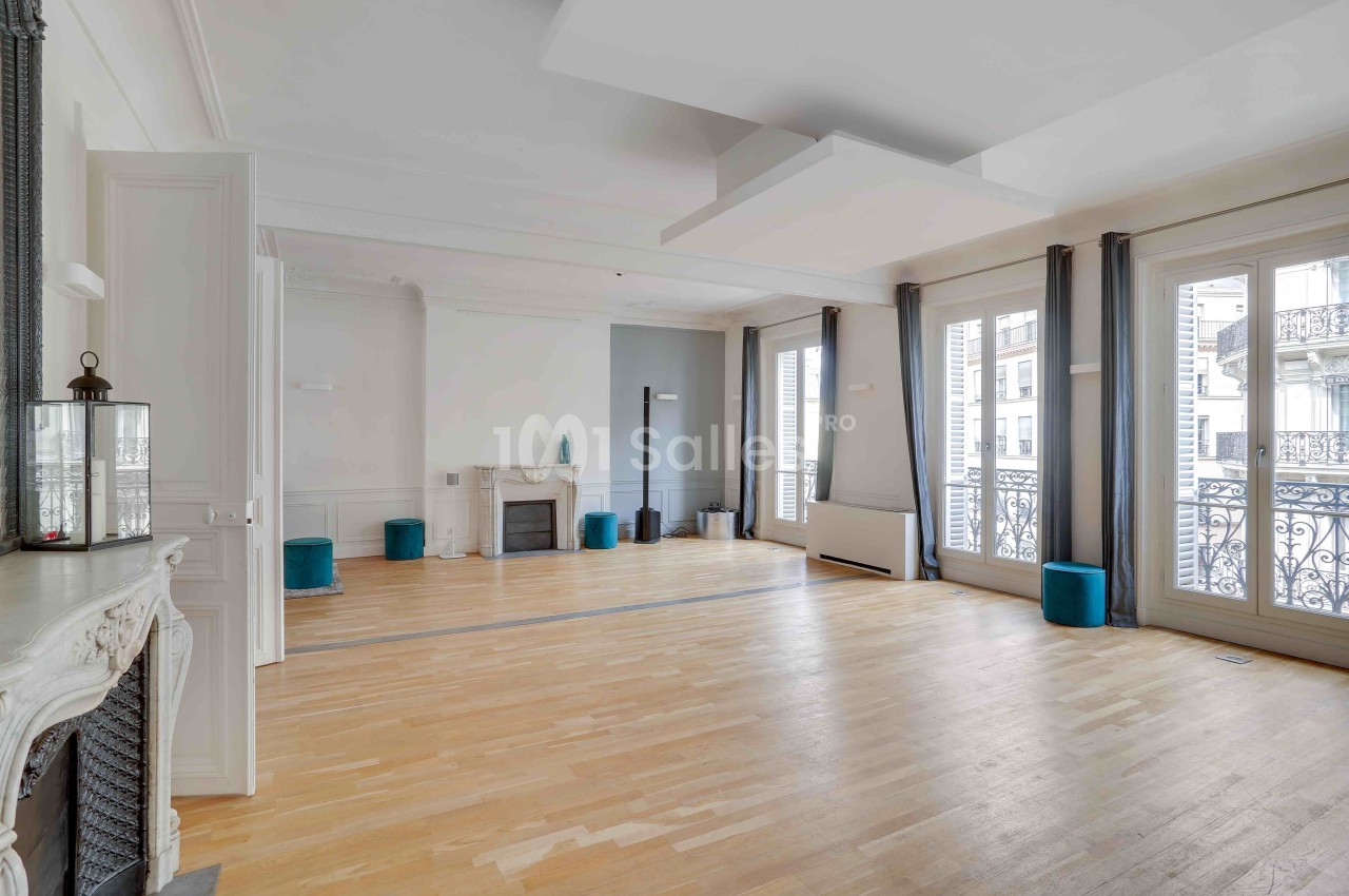 Salle lumineuse avec parquet clair, cheminée décorative, grandes fenêtres et rideaux gris donnant sur des balcons en fer…
