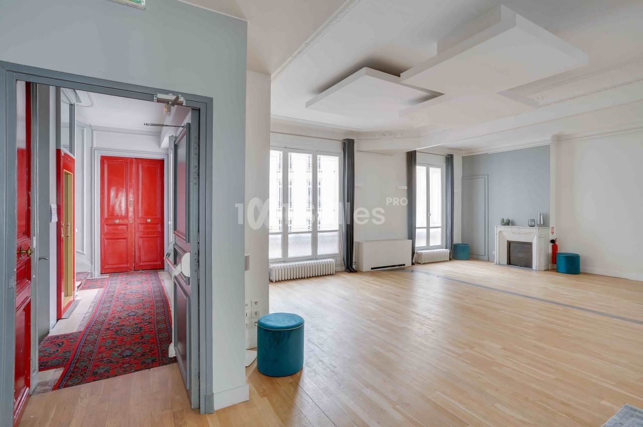 Salle lumineuse avec parquet clair, grandes fenêtres, plafonniers modernes et portes rouges visibles dans une autre pièce.