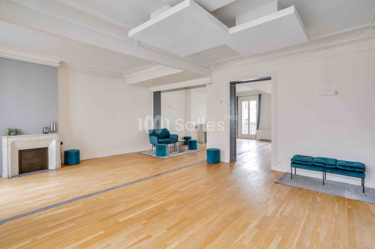 Salon lumineux avec parquet clair, cheminée décorative, murs blancs, fauteuils bleu-vert et moulures au plafond.