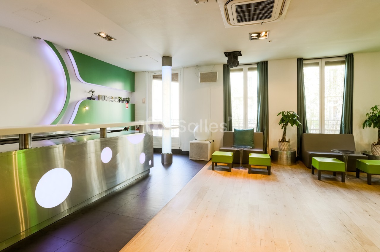 Intérieur lumineux avec comptoir moderne, sièges verts, plantes et grandes fenêtres laissant entrer la lumière naturelle.