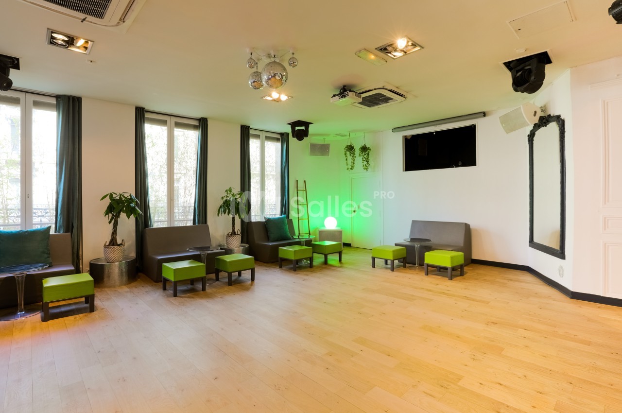 Salle lumineuse avec parquet clair, sièges gris et verts, grandes fenêtres, plantes et éclairage décoratif.