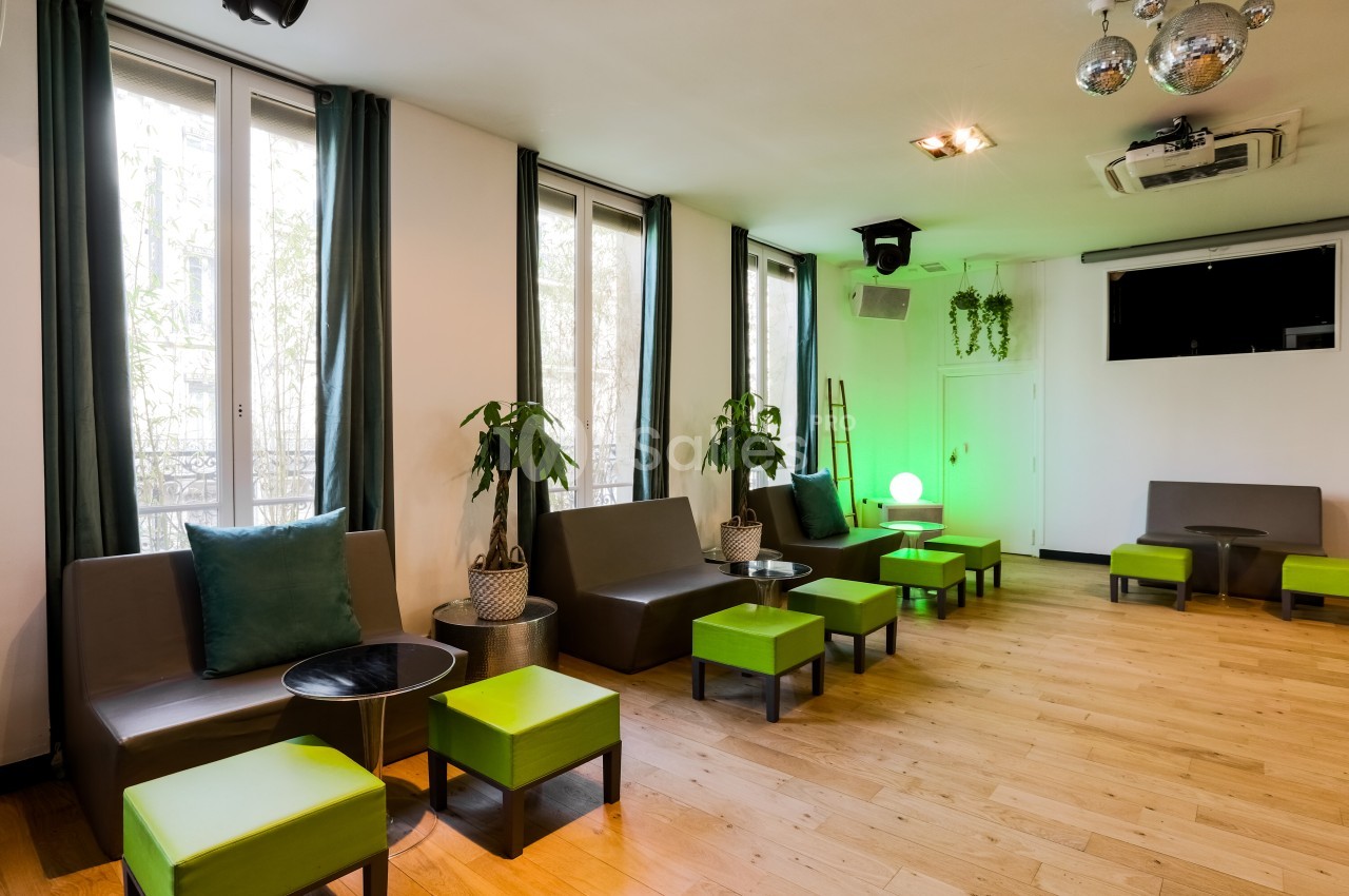 Salle lumineuse avec parquet, banquettes grises, poufs verts, plantes et décoration moderne, éclairée par des fenêtres.