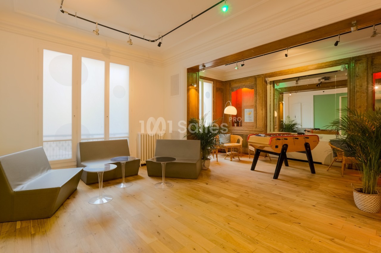 Salle lumineuse avec parquet, canapés gris, baby-foot, plantes et grandes fenêtres laissant entrer la lumière naturelle.