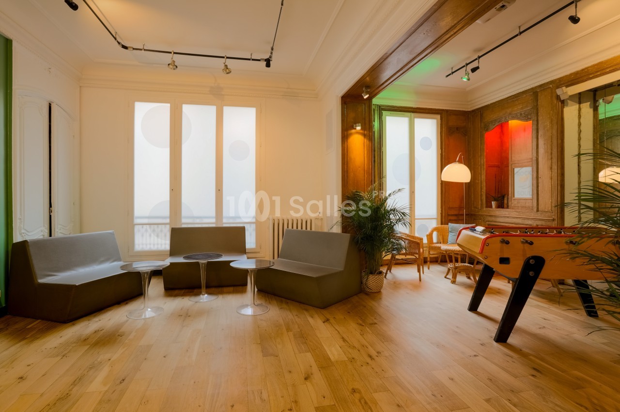 Espace intérieur lumineux avec parquet, canapés, baby-foot, plantes et grandes fenêtres.