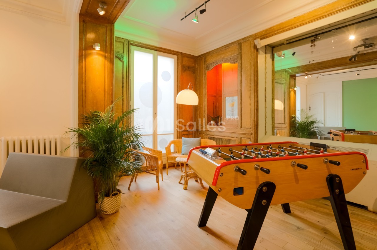 Salle lumineuse avec baby-foot, fauteuils en rotin, plantes et murs en bois, ambiance conviviale.