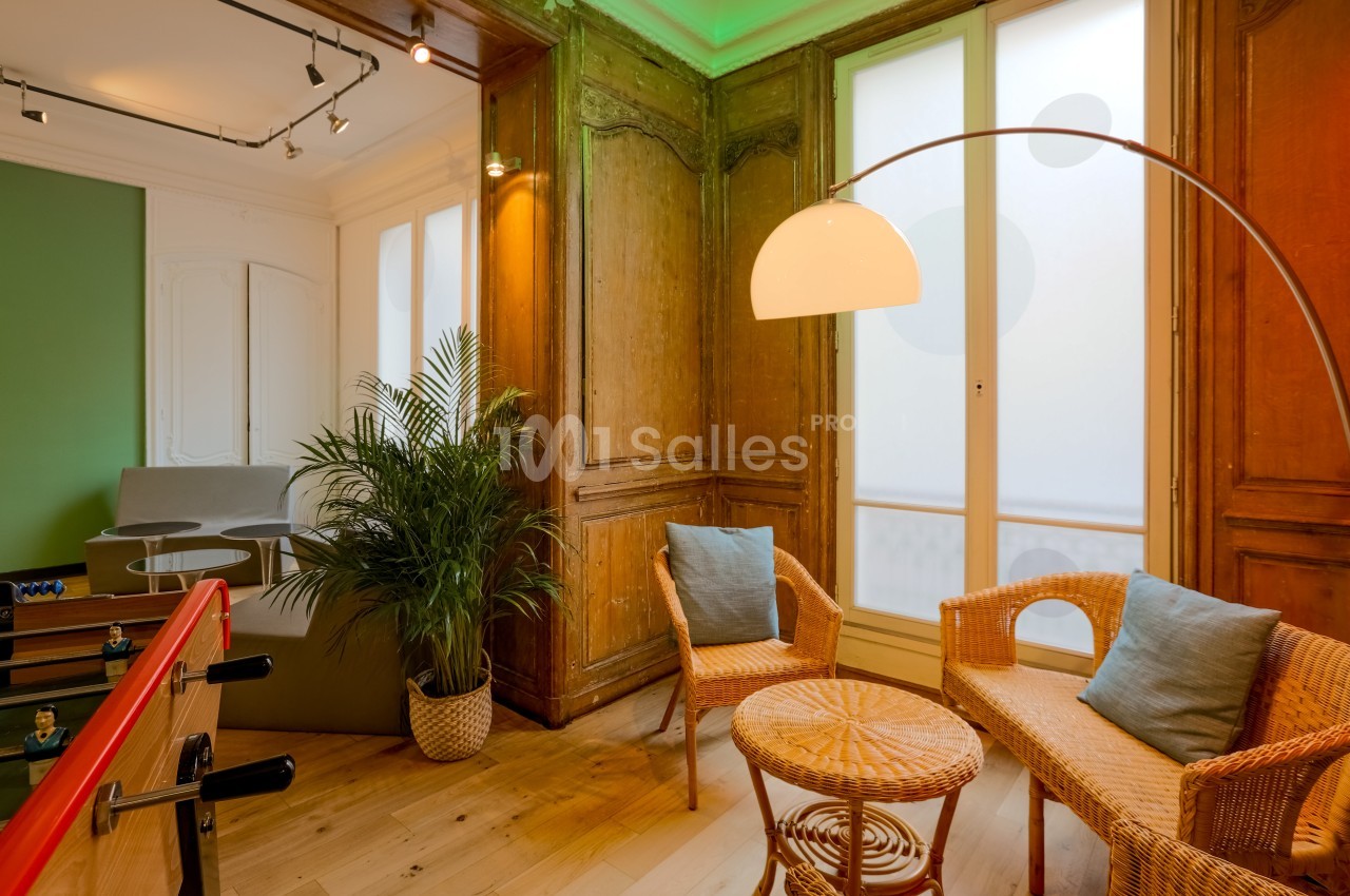 Espace lumineux avec fauteuils en rotin, table basse, plante verte et baby-foot, encadré de boiseries et grandes fenêtres.