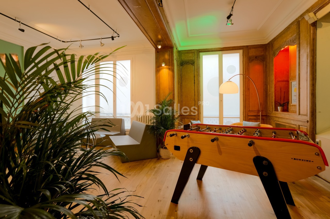 Salle lumineuse avec baby-foot en bois, fauteuils modernes et plantes, décorée de boiseries et éclairée par une grande…