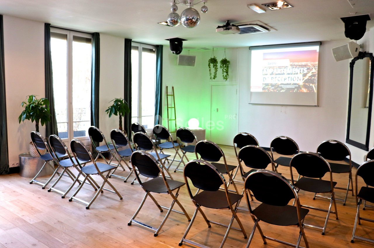 Salle lumineuse avec chaises pliantes disposées en rangées face à un écran de projection, plantes et décor sobre.