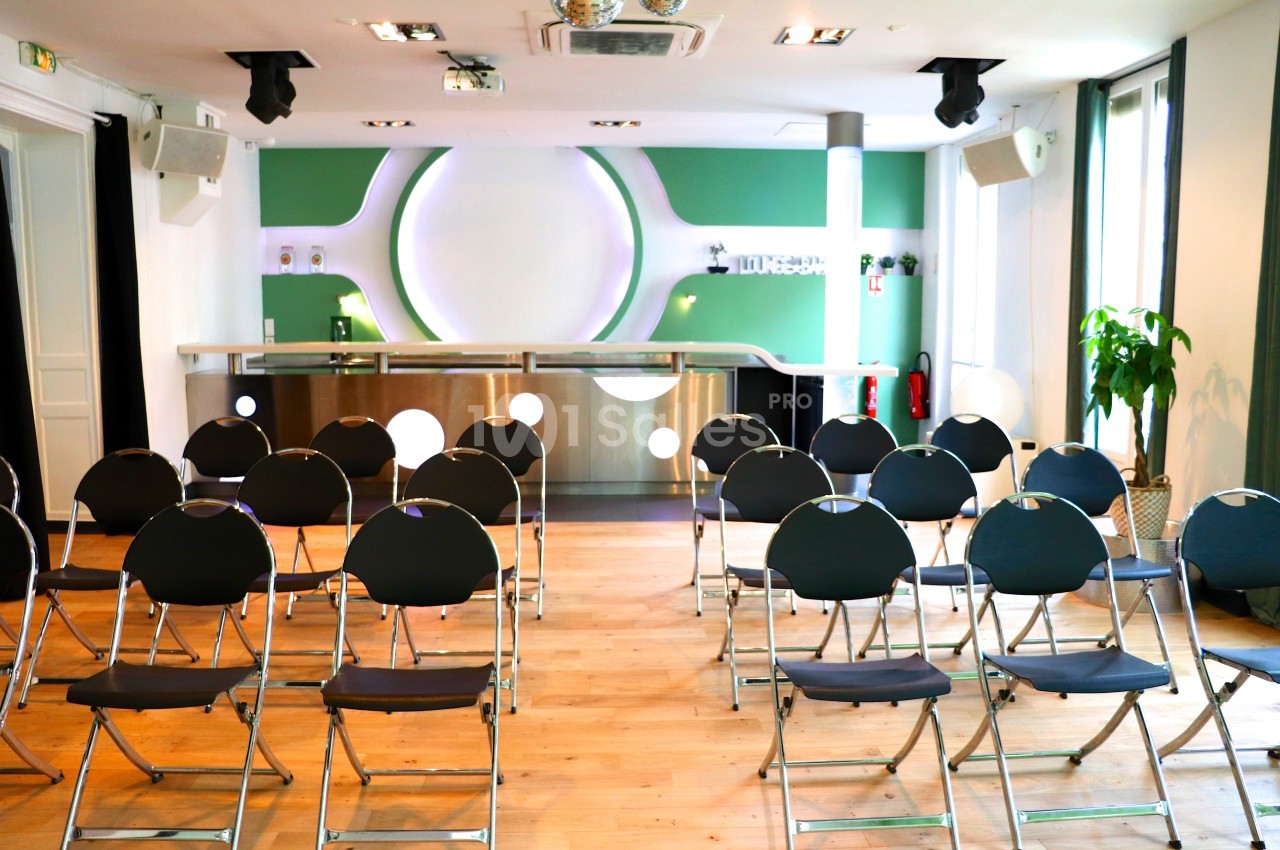 Salle lumineuse avec chaises pliantes disposées face à un comptoir moderne, murs verts et éclairage circulaire.