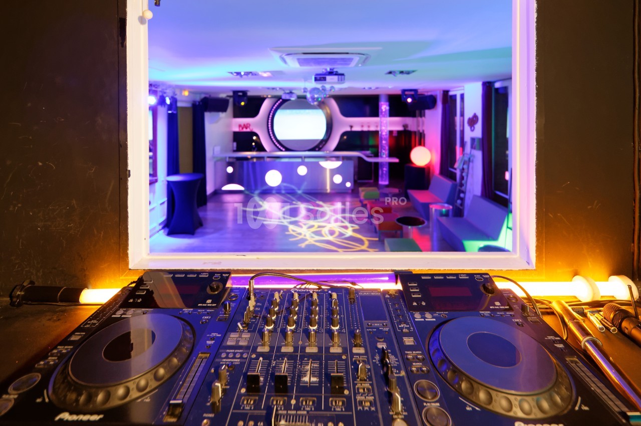 Vue d'une table de mixage DJ en premier plan avec une salle colorée et éclairée en arrière-plan.