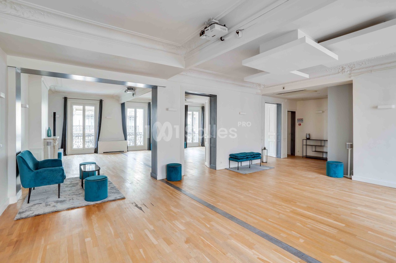 Salle spacieuse et lumineuse avec parquet, fauteuils bleus, grandes fenêtres et éléments décoratifs modernes.