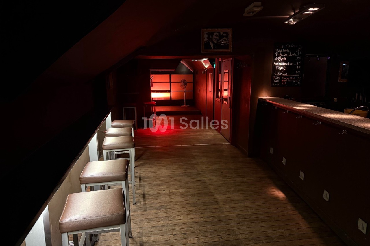 Salle sombre avec un éclairage rouge au fond, tabourets alignés à gauche et un comptoir à droite.