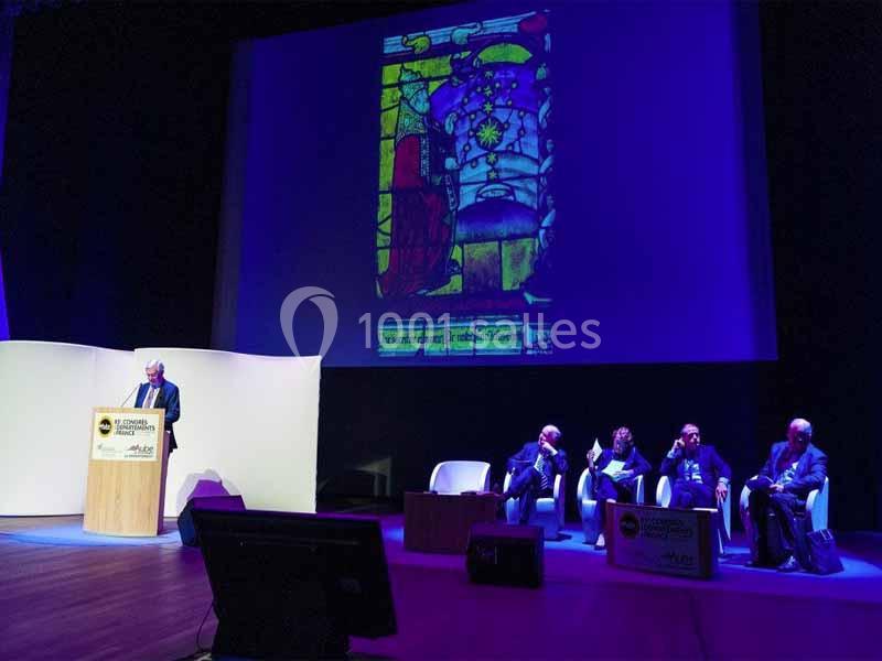 Un conférencier s'exprime sur scène devant un public, avec une image de vitrail projetée en arrière-plan.