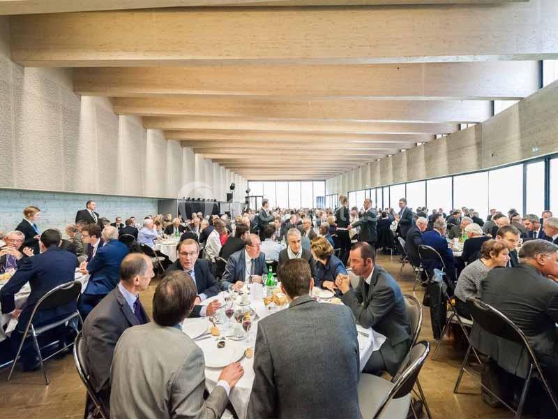Des personnes assises à des tables rondes participent à un événement dans une grande salle lumineuse avec un plafond en bois.
