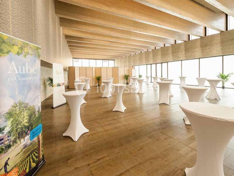 Salle lumineuse avec de grandes baies vitrées, tables hautes recouvertes de nappes blanches et un panneau promotionnel.