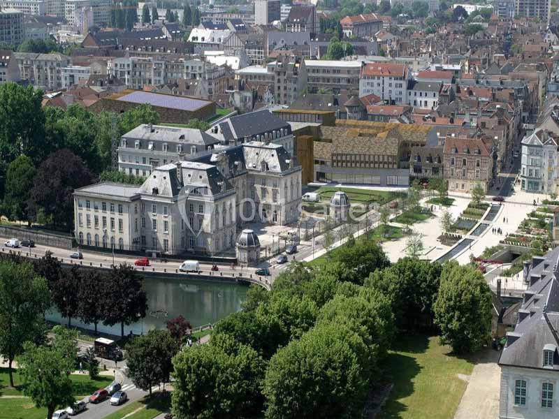 Vue aérienne d'un quartier urbain avec un bâtiment historique, un parc arboré et une rivière bordée d'arbres.