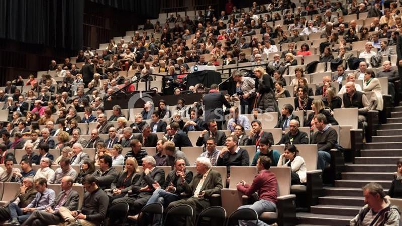 Salle comble avec des spectateurs assis dans un amphithéâtre, attentifs à un événement ou une présentation.