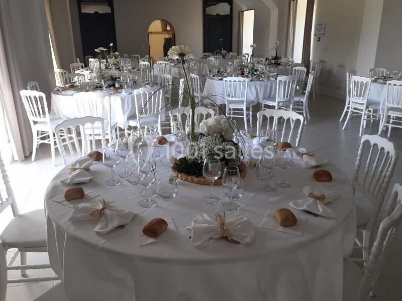 Salle de réception avec tables rondes dressées, nappes blanches, verres, serviettes pliées et décorations florales.
