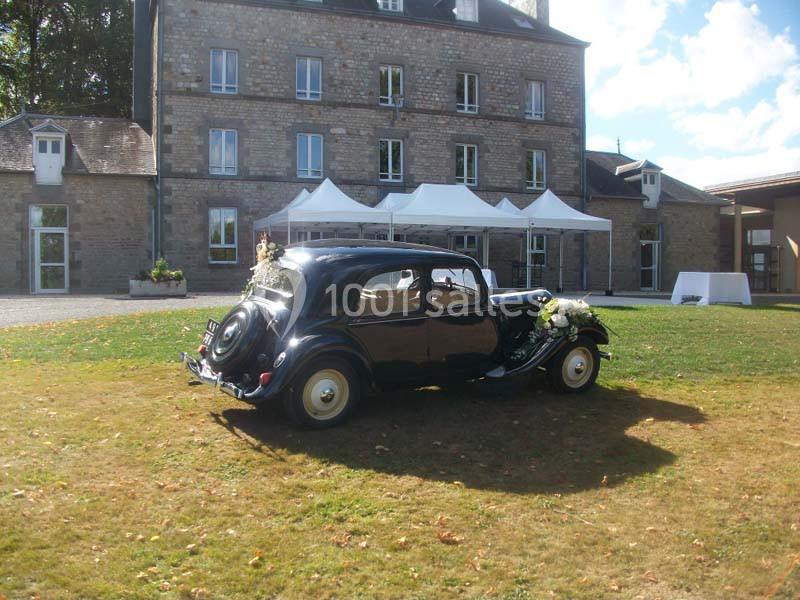 Voiture ancienne noire décorée pour un mariage, stationnée sur une pelouse devant un bâtiment en pierre.