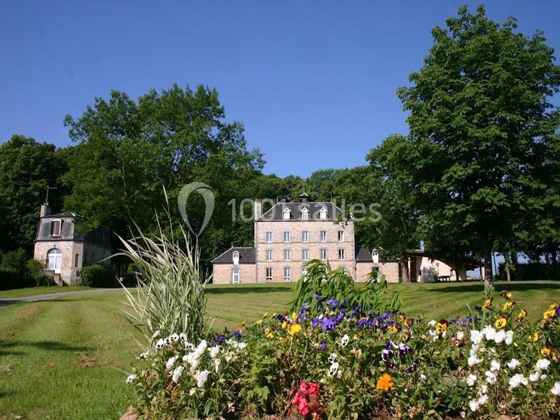 Manoir en pierre entouré de pelouses, arbres et parterre de fleurs colorées sous un ciel bleu.