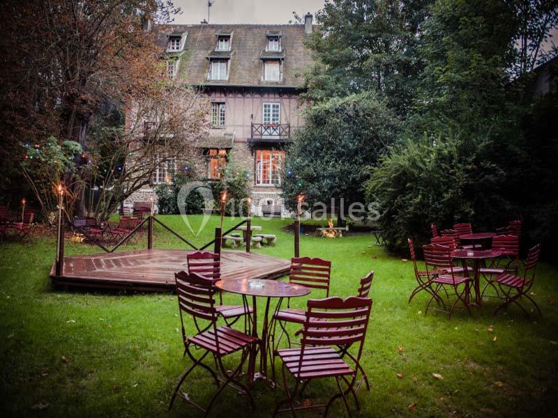 Jardin avec pelouse, tables et chaises rouges, scène en bois, entouré d'arbres et d'une maison à colombages.