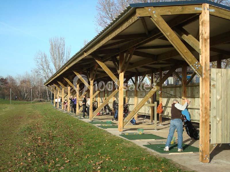 Des personnes s'entraînent au golf sous un abri en bois, alignées sur des postes de practice en extérieur.