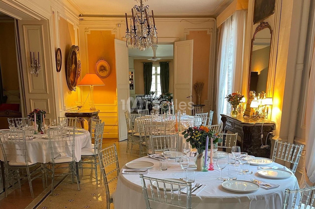 Salle élégante avec tables rondes dressées, chaises transparentes, lustre, miroirs et lumière naturelle.