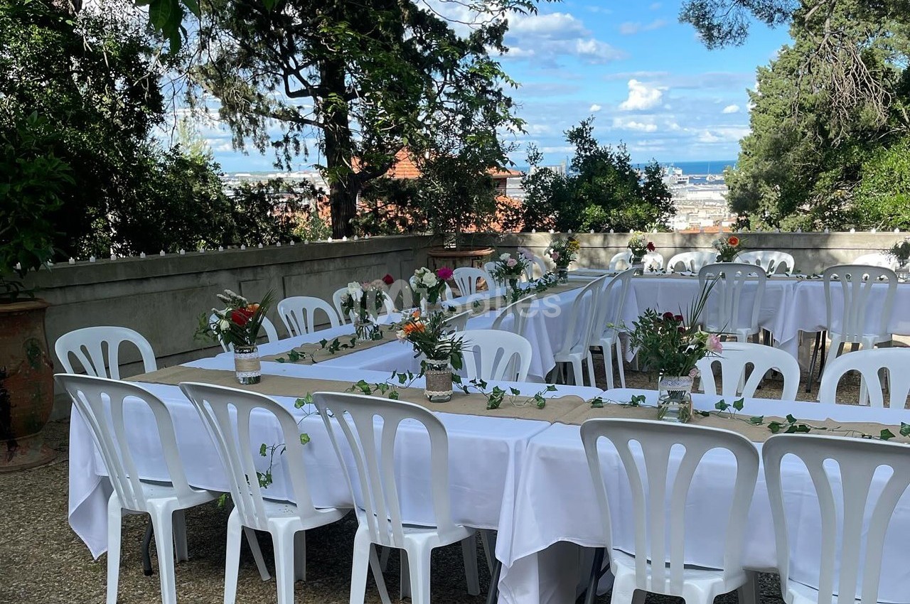 Tables blanches décorées de fleurs et chaises en plastique disposées en extérieur avec vue sur un paysage urbain.