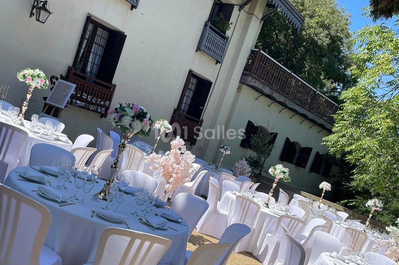 Tables rondes décorées pour un événement en plein air, disposées devant un bâtiment avec balcons et volets en bois.