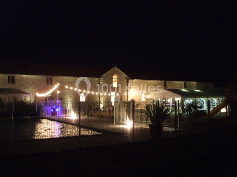 Bâtiment en pierre éclairé la nuit, avec guirlandes lumineuses et piscine entourée d'une clôture au premier plan.