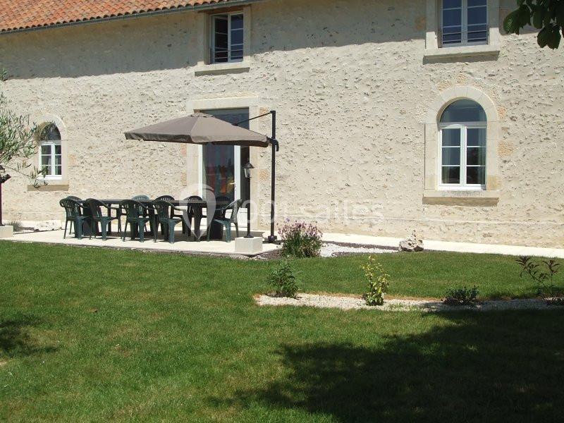 Terrasse avec table, chaises en plastique et parasol, située devant une maison en pierre avec jardin gazonné.