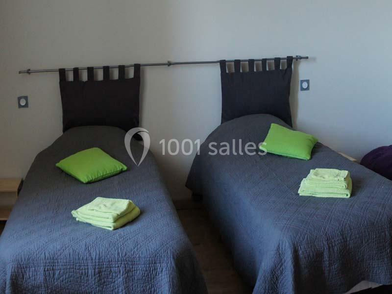 Deux lits simples avec couvre-lits gris, oreillers verts et serviettes pliées sur chacun, dans une chambre sobre.