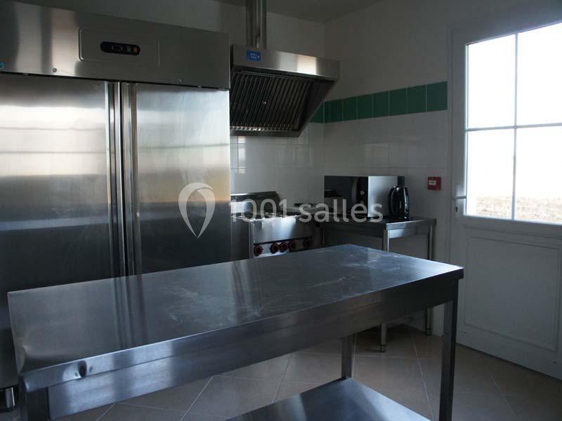 Cuisine professionnelle équipée avec réfrigérateur inox, hotte, plan de travail métallique et micro-ondes près d'une fenêtre.