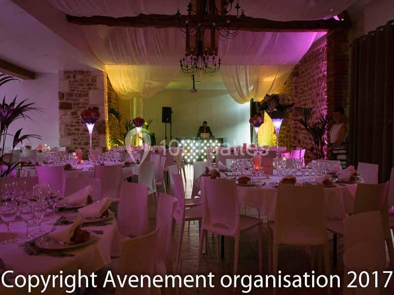 Salle de réception décorée avec des tables dressées, éclairage coloré et un DJ au fond près d'une console lumineuse.