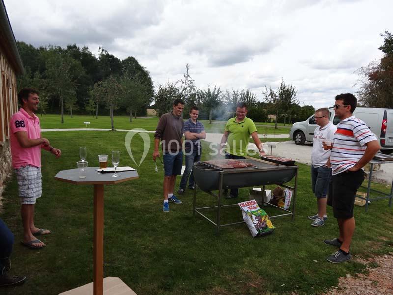 Des personnes discutent autour d'un barbecue allumé dans un jardin, avec des arbres et une pelouse en arrière-plan.