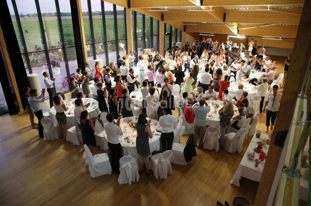 Salle lumineuse avec de grandes baies vitrées, remplie de convives debout et assis autour de tables décorées.