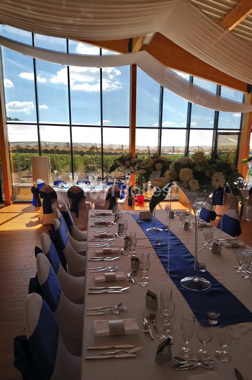 Salle de réception lumineuse avec tables décorées de nappes blanches et bleues, et vue sur un paysage extérieur ensoleillé.