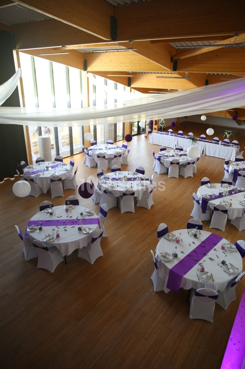 Salle de réception décorée avec des tables rondes dressées, nappes blanches et chemins de table violets.