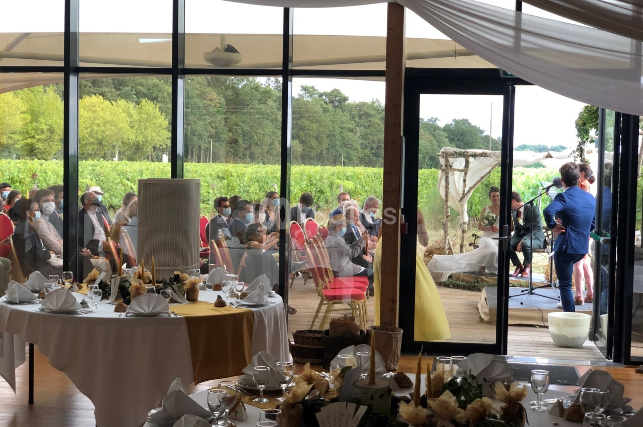 Des invités assis assistent à une cérémonie en intérieur avec vue sur des vignes à travers de grandes baies vitrées.