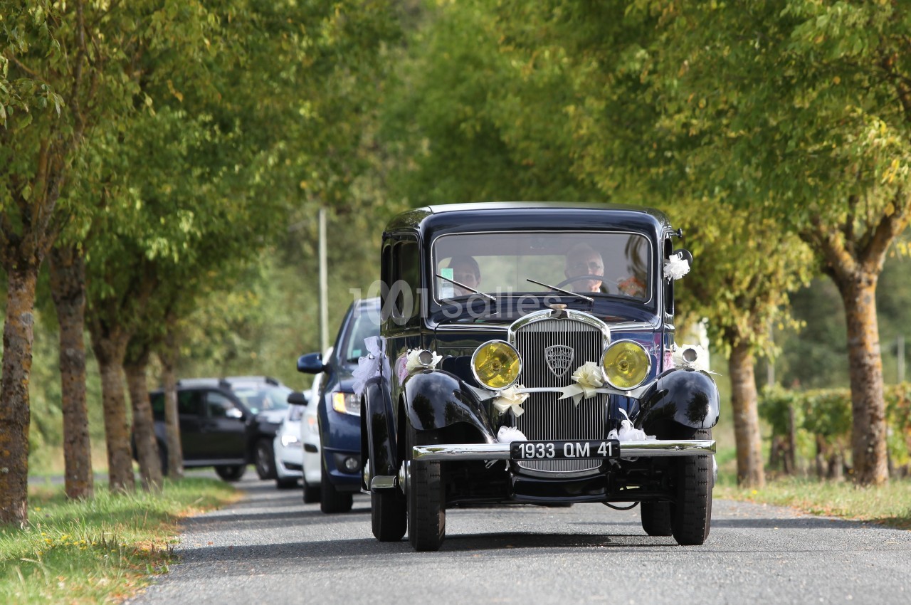 Voiture ancienne décorée pour un mariage roulant sur une route bordée d'arbres, suivie par d'autres véhicules.
