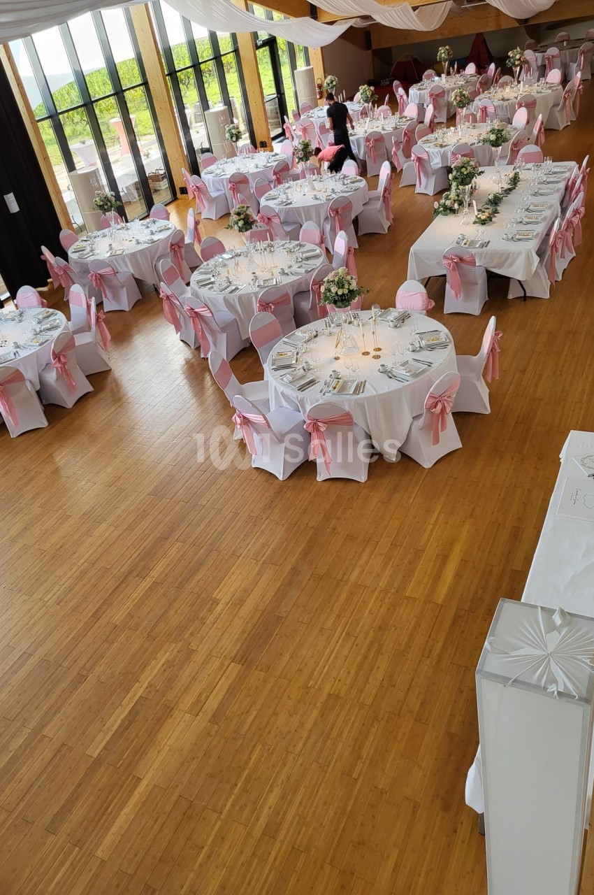 Salle de réception décorée avec des tables rondes, nappes blanches et chaises ornées de nœuds roses.