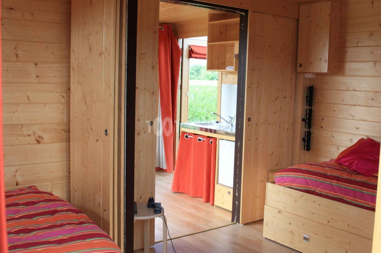 Intérieur d'un hébergement en bois avec deux lits, une kitchenette et des rideaux rouges donnant sur l'extérieur.