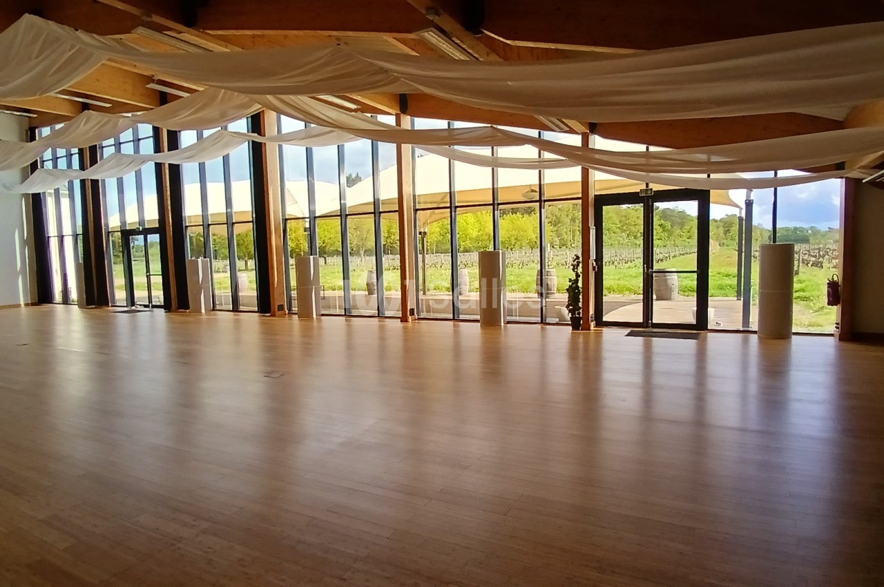 Salle lumineuse avec parquet en bois, grandes baies vitrées et rideaux blancs, donnant sur un espace extérieur verdoyant.