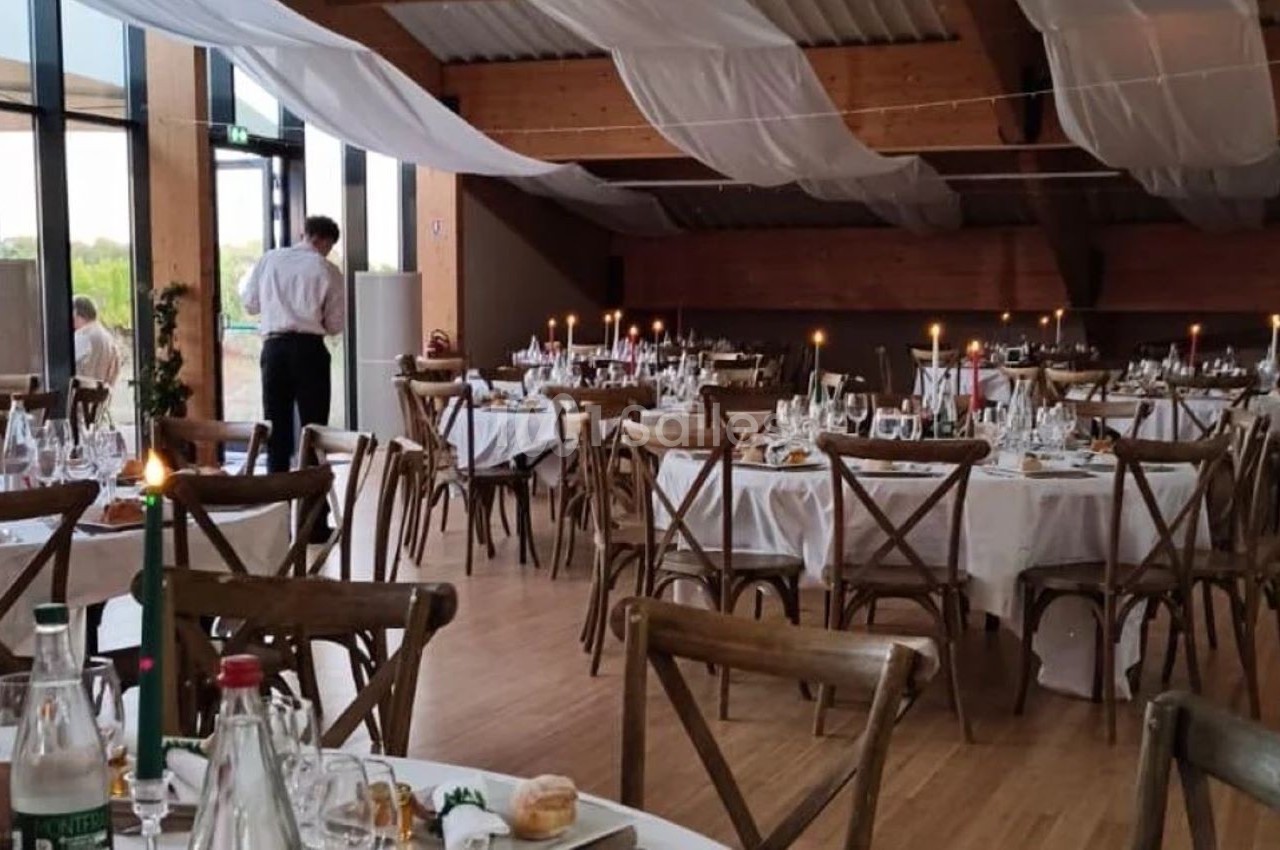Salle de réception décorée avec des tables rondes dressées, bougies allumées et nappes blanches, sous un plafond en bois.