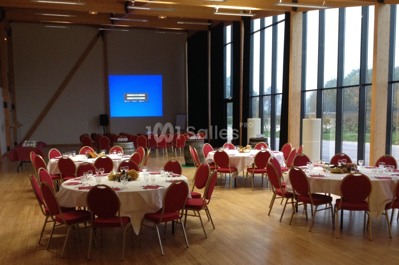Salle de réception lumineuse avec des tables rondes dressées et des chaises rouges, prête pour un événement.