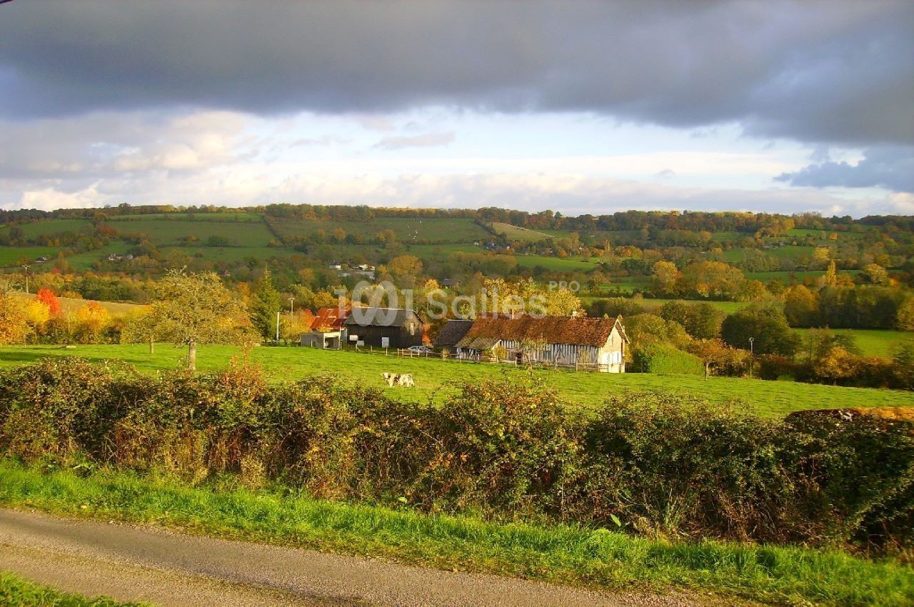 Paysage rural avec une ferme traditionnelle entourée de prairies verdoyantes et de collines sous un ciel nuageux.