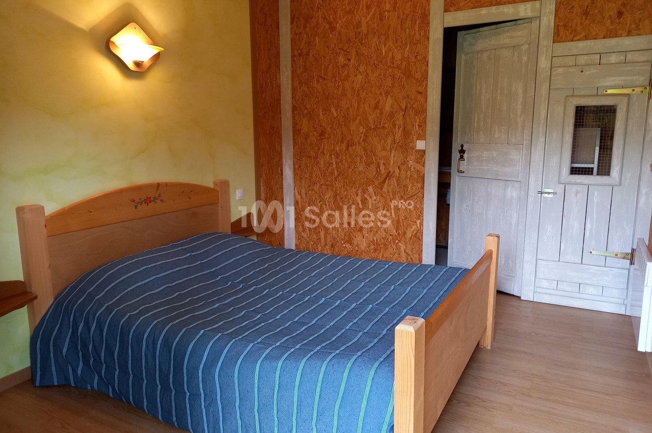 Chambre avec un lit double en bois, couverture bleue rayée, murs en bois et plâtre, éclairage mural allumé.