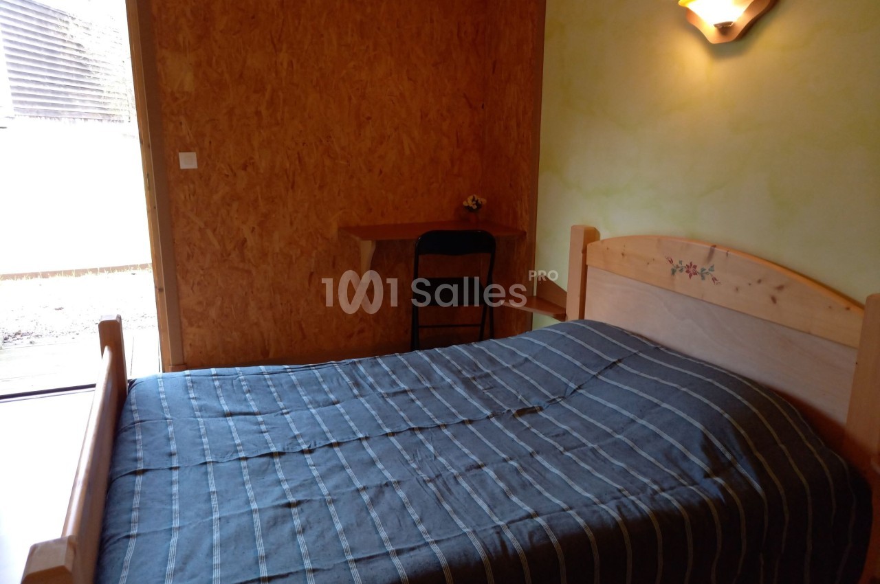 Chambre simple avec un lit double, une table en bois, une chaise et une lumière murale, donnant sur une porte vitrée.
